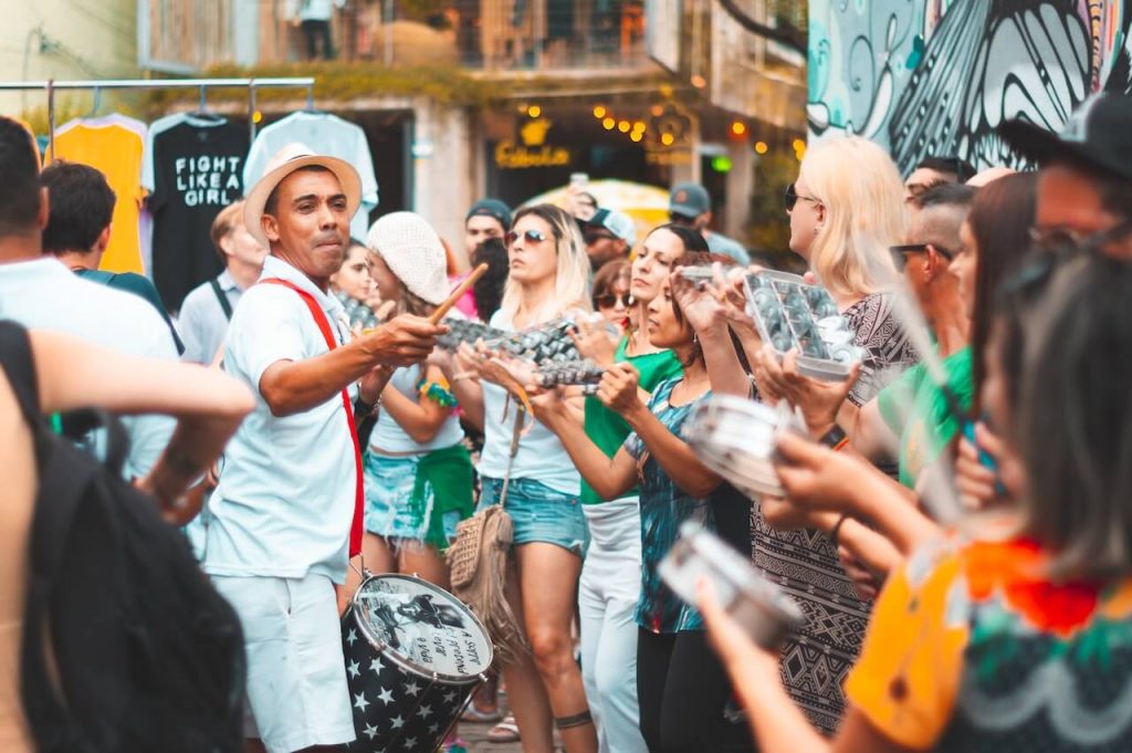 Imagem de um grupo de pessoas nas ruas tocando instrumentos de samba com animação e alegria. 